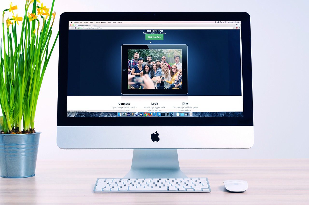 Building a website : This image shows a computer screen and keyboard on a light oak desk. The screen shows a website image of a group of friends with navigation options below it. To the left of the screen there is a grey plant pot with daffodils growing in it.